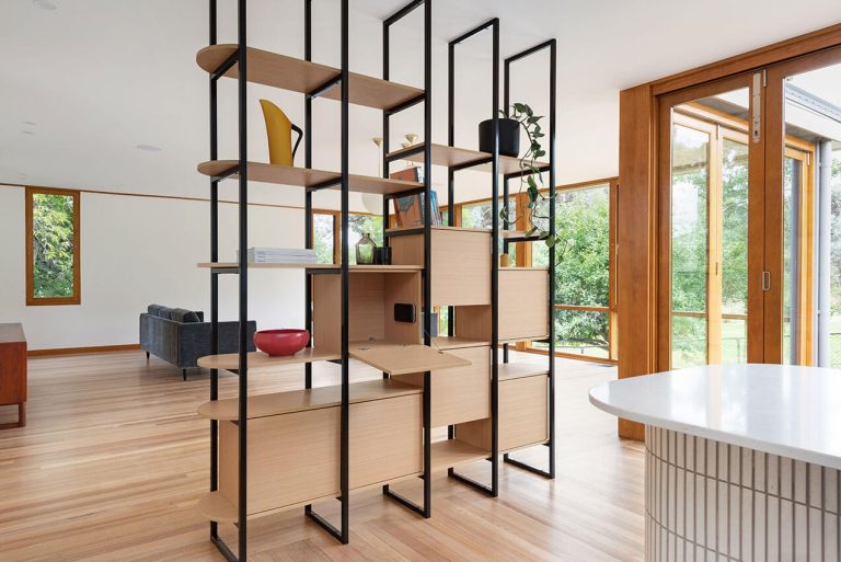 Image shows the interior of a modernist house, with a see-through bookshelf separating a lounge room and kitchen area. There are large timber windows looking out over a garden.