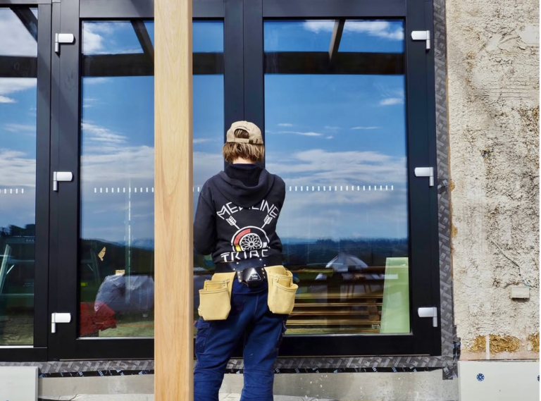 Image shows the exterior of a 9 star house in Tasmania with rendered strawbale walls and a person standing in front of large triple-glazed windows.